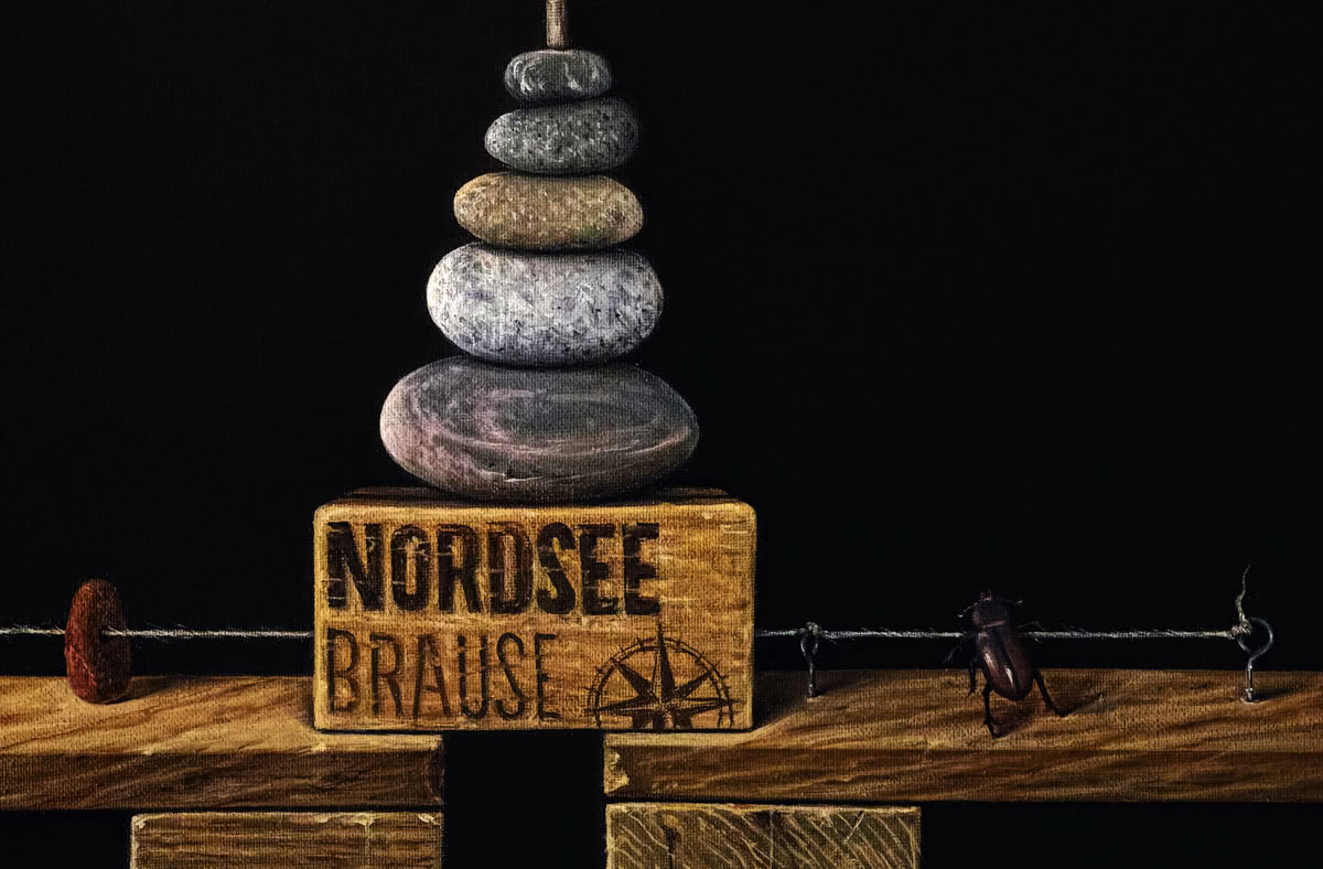 Ein Stapel aus sieben Steinen auf einem Holzblock mit der Aufschrift "Nordsee Brause", vor dunklem Hintergrund (Detail).